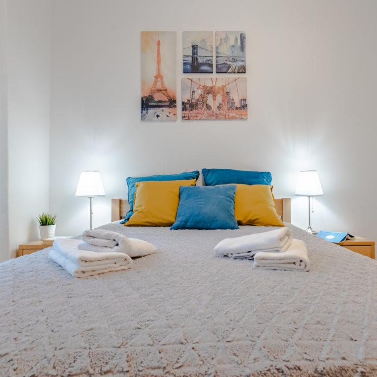 Jasna sypialnia z łóżkiem, niebiesko-żółtymi poduszkami, lampkami nocnymi i obrazami wieżowców na ścianie.