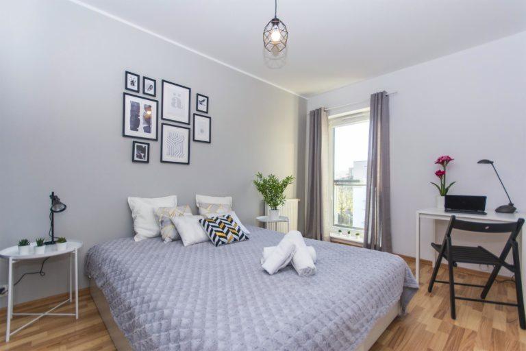 Jasna sypialnia z łóżkiem, galerią obrazków na szarej ścianie, biurkiem z laptopem i kwiatami, oraz lampą wiszącą w kształcie geometrycznej klatki.