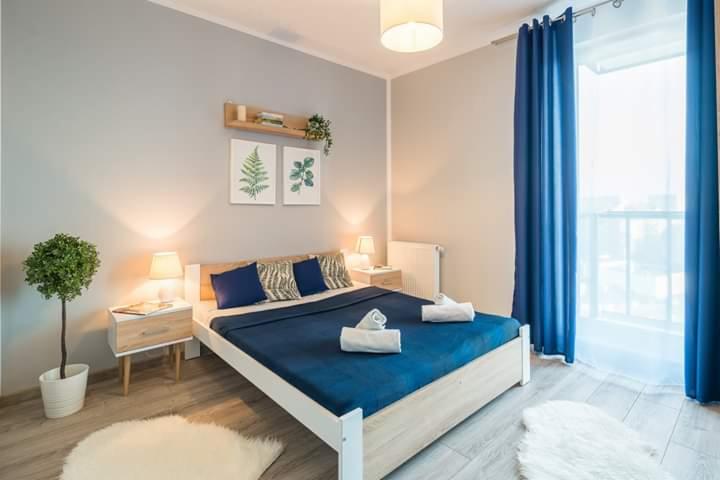 Jasna sypialnia z łóżkiem z niebieską narzutą, białymi ręcznikami, drewnianą podłogą i dekoracjami roślinnymi na ścianie oraz niebieskimi zasłonami w oknie.