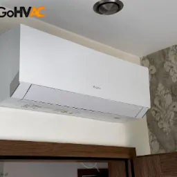 Biały klimatyzator marki Gree zamontowany na ścianie z wzorzystą tapetą, widoczny nad drewnianą ramą drzwiową i fragmentem szafki, logo GoHVAC w lewym górnym rogu.