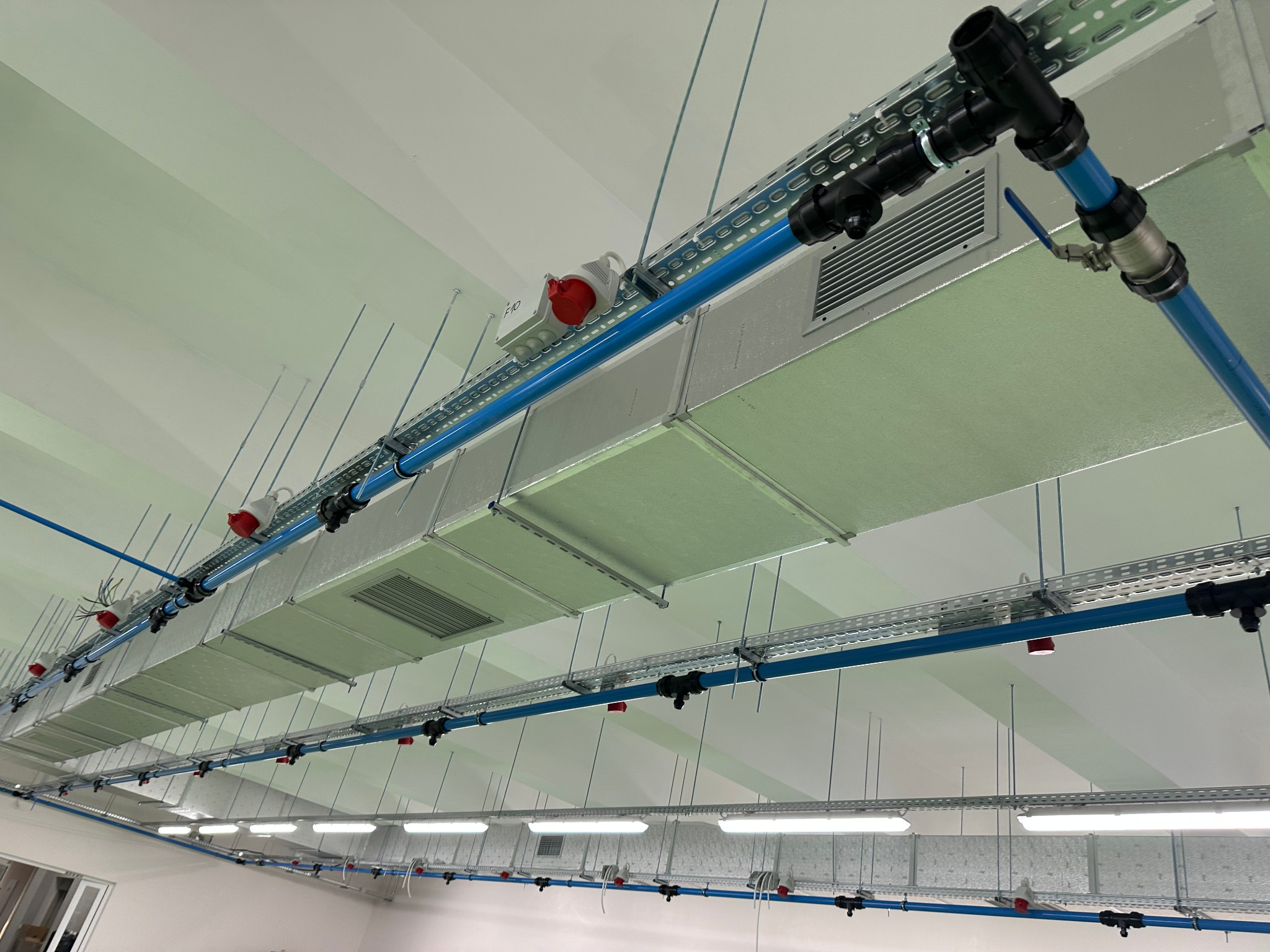 Kompletna instalacja wentylacji mechanicznej z izolowanymi kanałami PIR, niebieskimi rurami i czerwonymi gniazdami, zamontowana pod sufitem w jasnym pomieszczeniu.