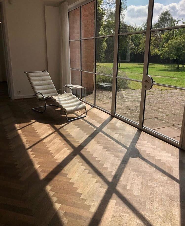 Parkiet w jodełkę w jasnym wnętrzu z leżanką i dużym oknem. Cienie okien tworzą geometryczne wzory na podłodze. Widok na zewnątrz z zielenią i błękitnym niebem.