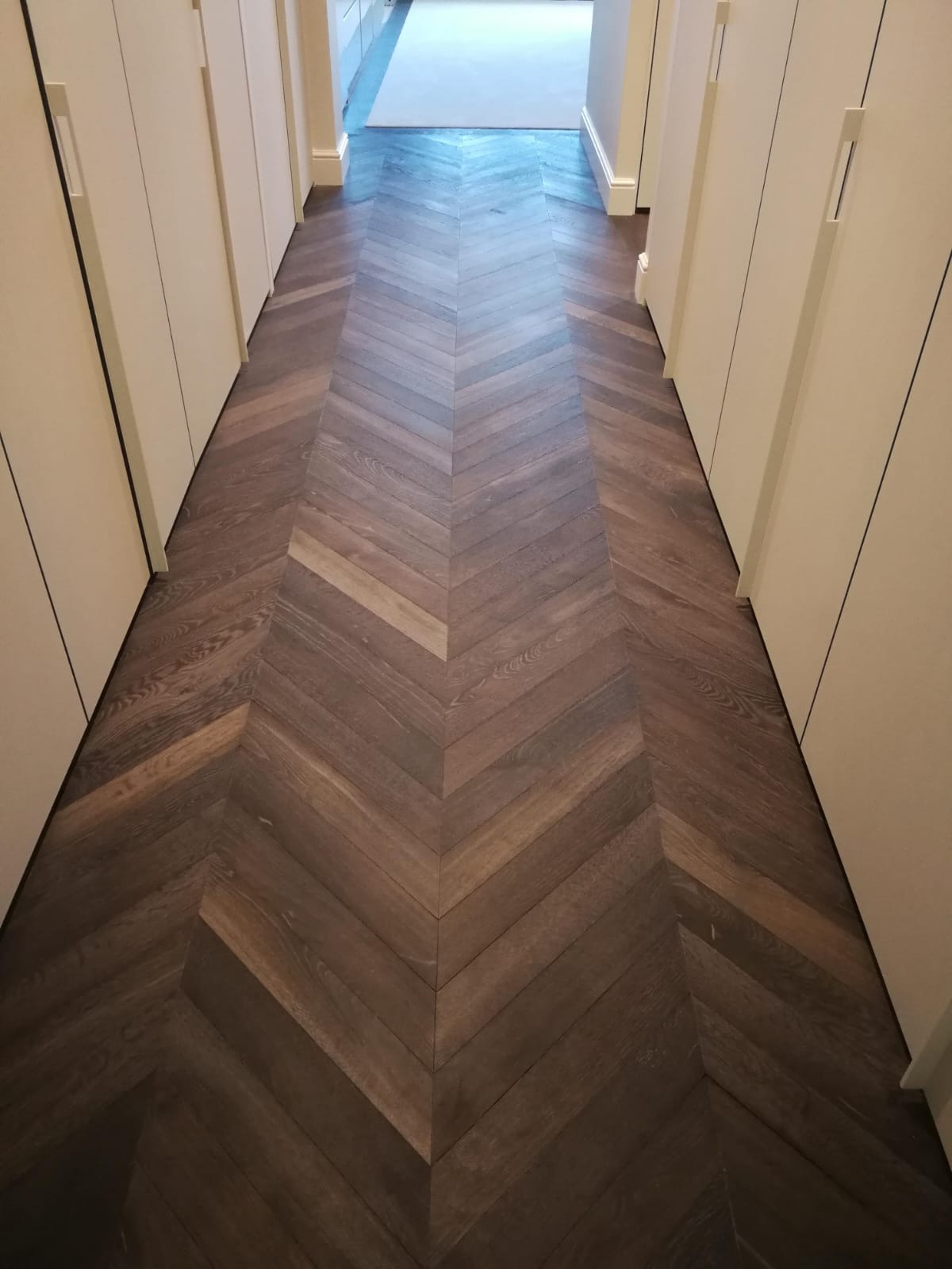 Ciemna, drewniana podłoga ułożona we wzór jodełki francuskiej w korytarzu z białymi szafami wnękowymi po obu stronach. Widok z góry na podłogę.
