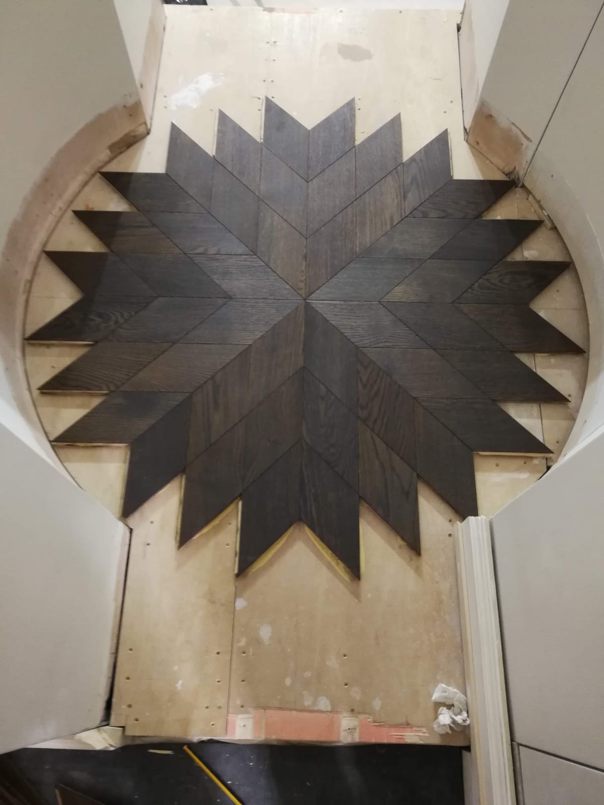 Układanie parkietu w kształcie gwiazdy z ciemnego drewna na podłodze z przygotowanym podłożem, widoczne fragmenty ścian i łukowate przejście.