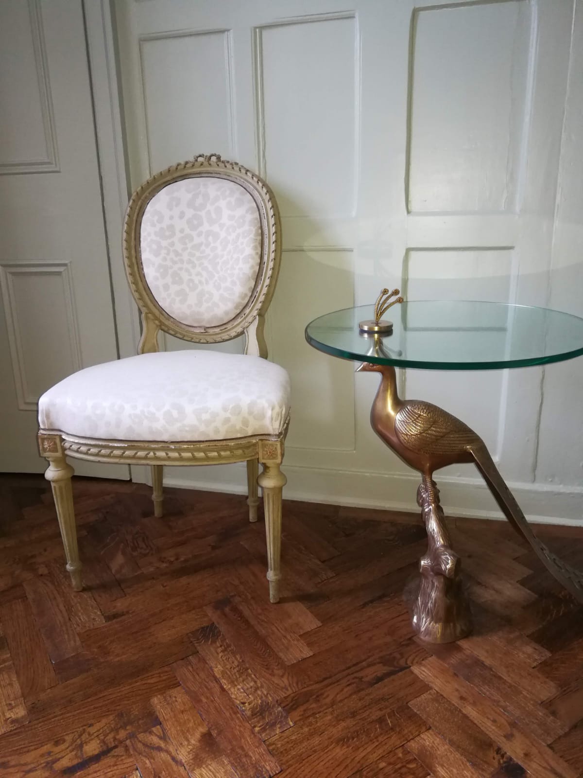 Stylowe krzesło z tapicerką w panterkę i stolik z podstawą w kształcie ptaka na parkiecie ułożonym we wzór jodełki, na tle panelowej ściany.