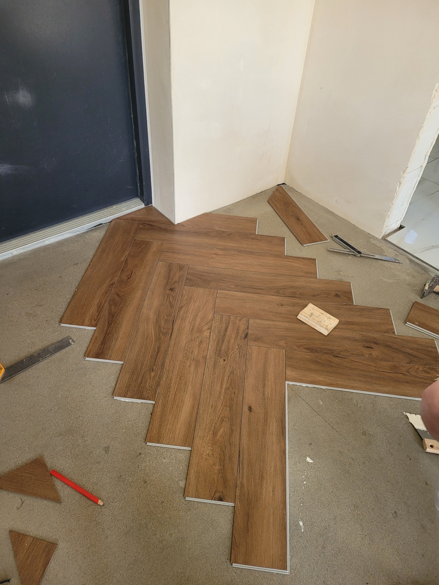 Proces układania paneli podłogowych w jodełkę, widoczne narzędzia i fragmenty paneli, pomieszczenie w trakcie remontu, surowy beton na podłodze.