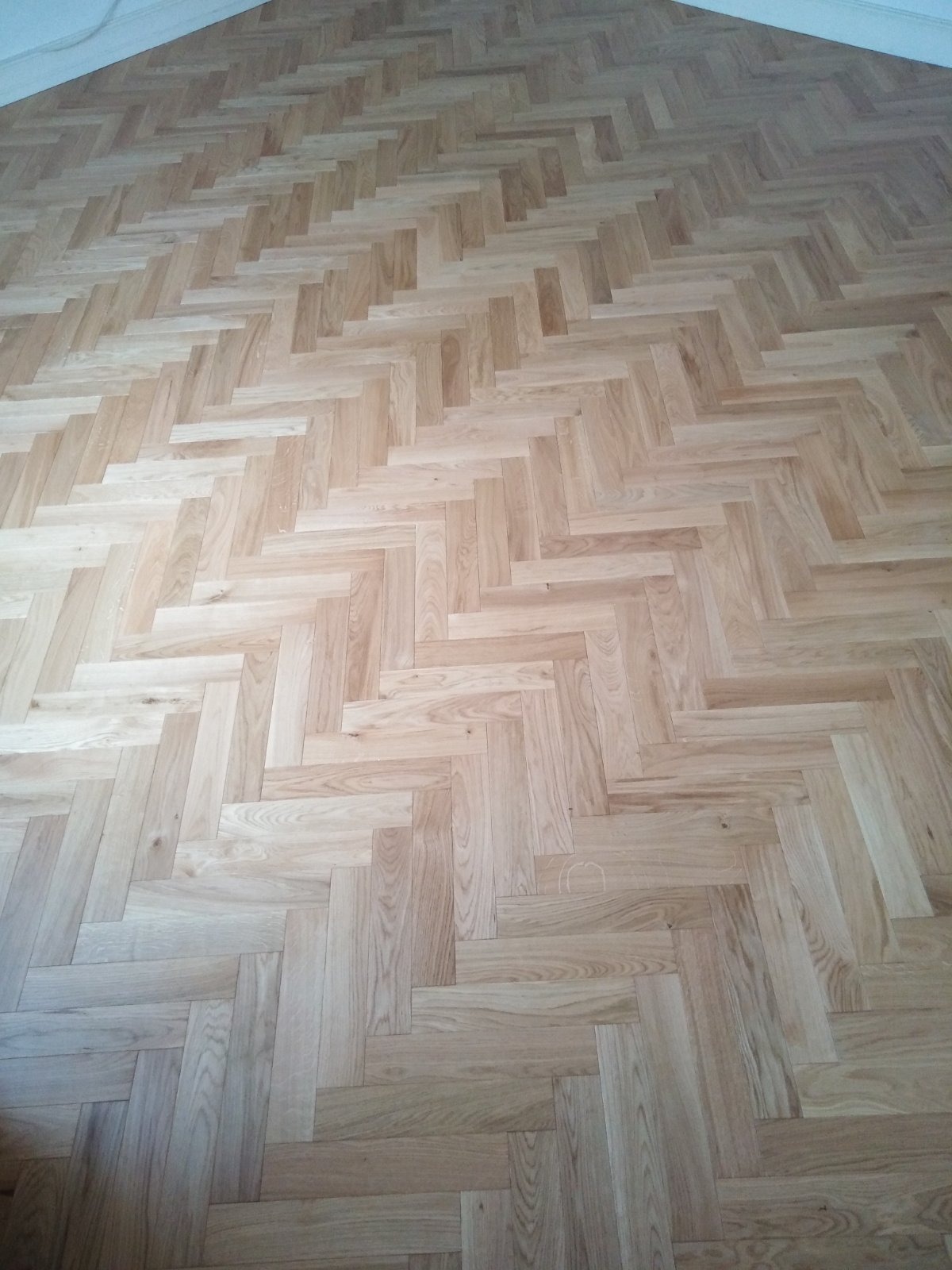 Jasny parkiet ułożony we wzór jodełki francuskiej, wypełniający kadr od podłogi do ściany. Widoczne naturalne usłojenie drewna i subtelne refleksy światła.