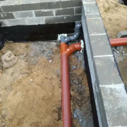 Ceglany fundament z hydroizolacją, widoczne rury kanalizacyjne w wykopie, szare rury połączone pianką montażową, ziemia i gruz w tle.