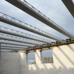 Wznoszenie ścian domu murowanego z betonu komórkowego, widoczne nadproża okienne i prefabrykowane belki stropowe zbrojone stalą, w tle pochmurne niebo.