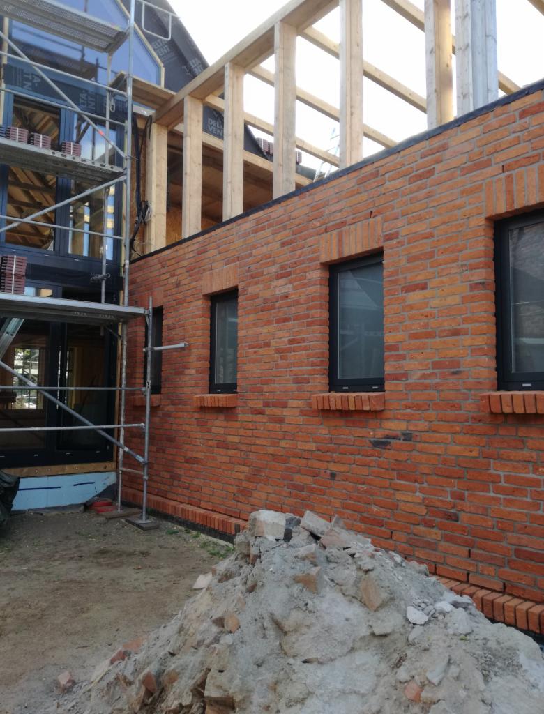 Wznoszenie ściany z czerwonej cegły z czarnymi oknami w trakcie budowy domu, widoczne rusztowanie i drewniana konstrukcja dachu.