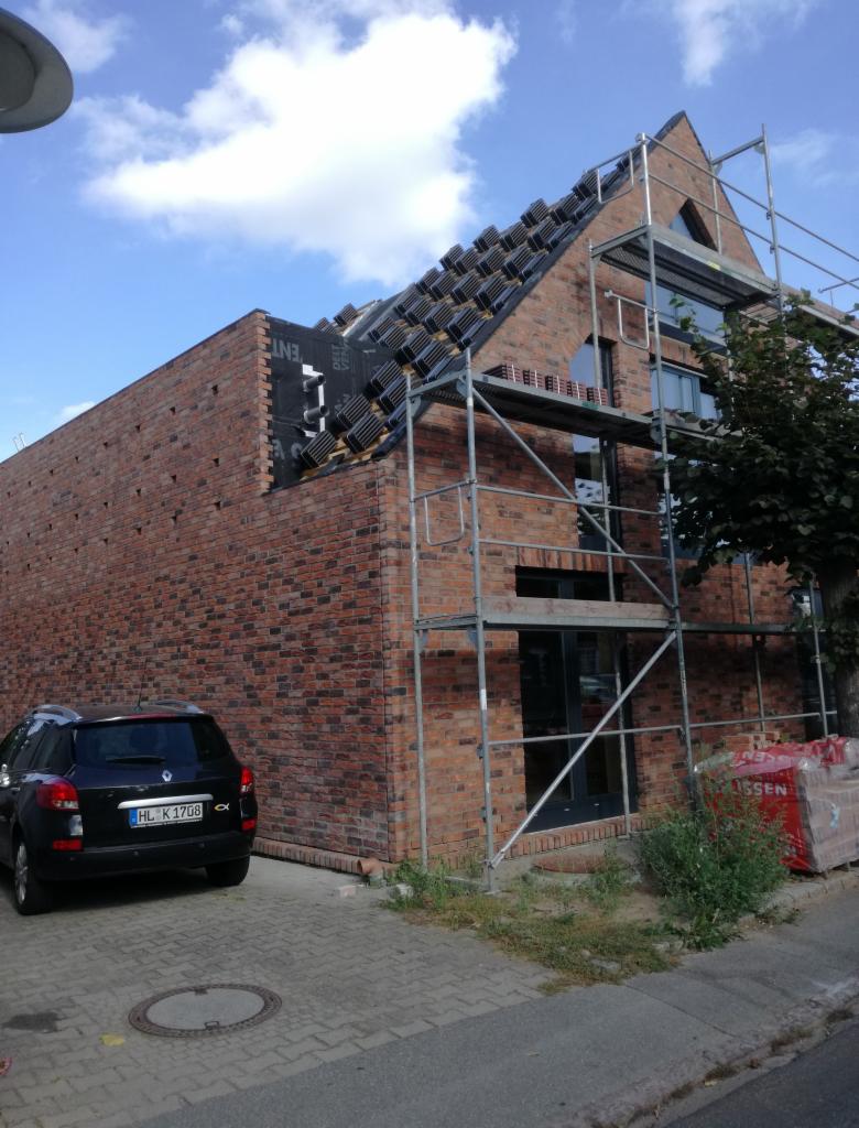 Wznoszony dom z czerwonej cegły, z widocznymi rusztowaniami, częściowo pokryty dachówką, z zaparkowanym czarnym samochodem osobowym na kostce brukowej.