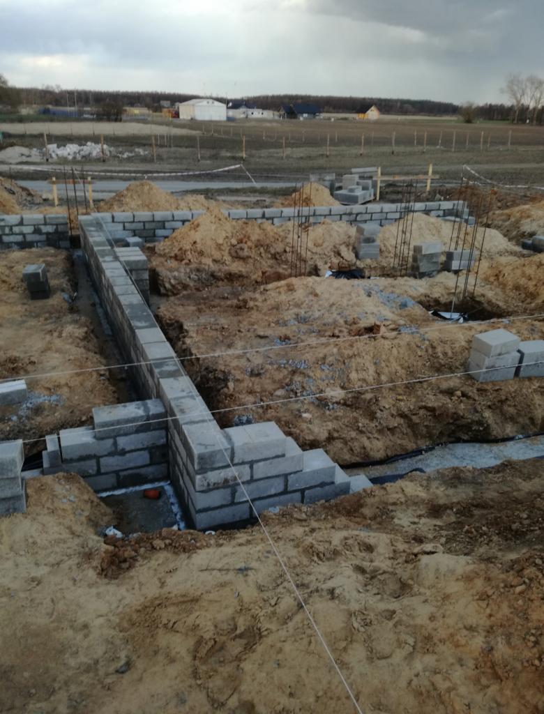 Wznoszenie fundamentów z bloczków betonowych na placu budowy, widoczne wystające pręty zbrojeniowe, ziemia i odległe budynki pod pochmurnym niebem.