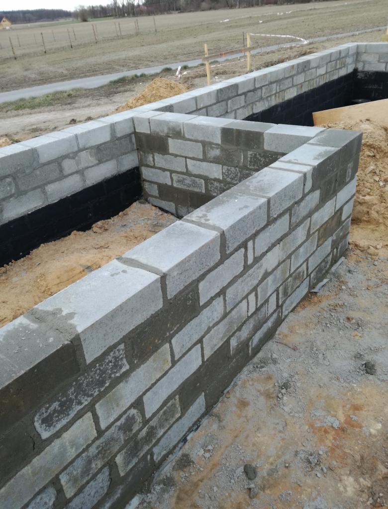 Wznoszenie ścian fundamentowych z bloczków betonowych, zabezpieczonych hydroizolacją, widok z góry na plac budowy.
