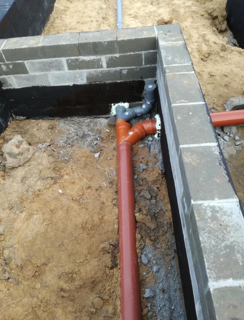 Ceglany fundament z hydroizolacją, widoczne rury kanalizacyjne w wykopie, szare rury połączone pianką montażową, ziemia i gruz w tle.