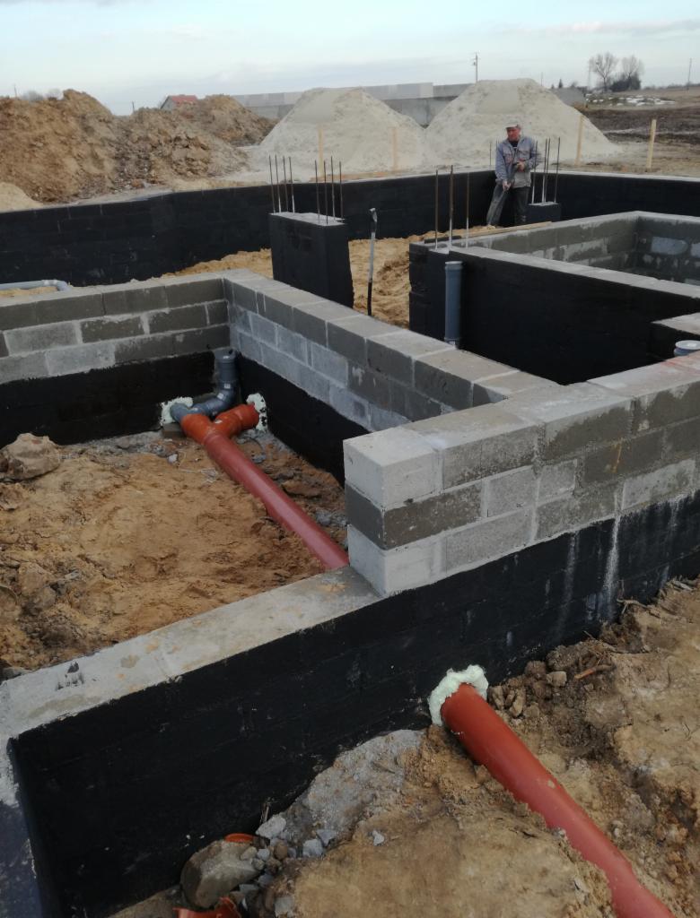 Wykop pod fundamenty z ułożonymi rurami kanalizacyjnymi, zaizolowane i wymurowane ściany fundamentowe z betonu komórkowego, widoczny pracownik w tle.