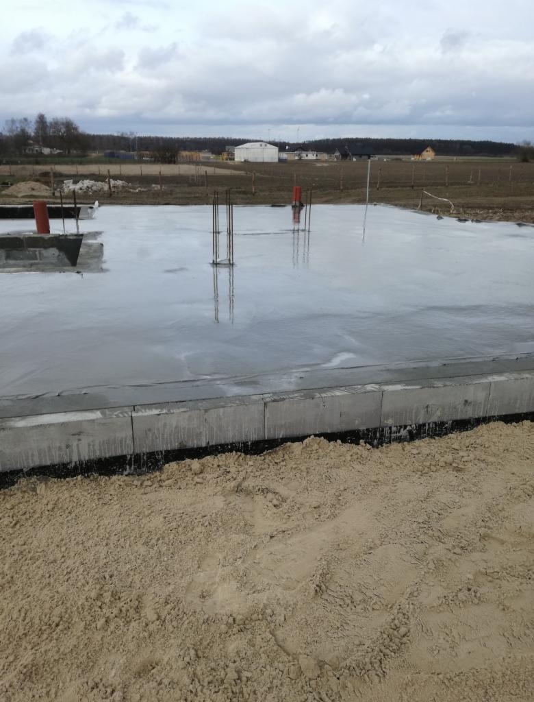Świeżo wylany fundament z wystającymi prętami zbrojeniowymi, widoczne bloczki fundamentowe i piasek zasypowy, pochmurne niebo w tle.