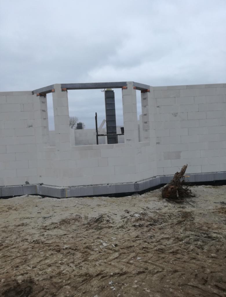 Wznoszone ściany z betonu komórkowego z widocznymi nadprożami w domu w budowie w Piechcinie, z korzeniem drzewa na pierwszym planie.