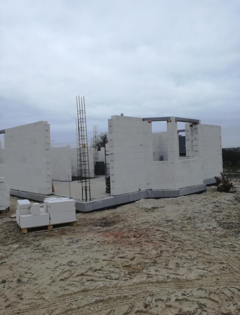 Wznoszenie ścian domu z białych bloczków na fundamencie, widoczne zbrojenia i paleta z bloczkami, piaszczyste podłoże, pochmurne niebo.