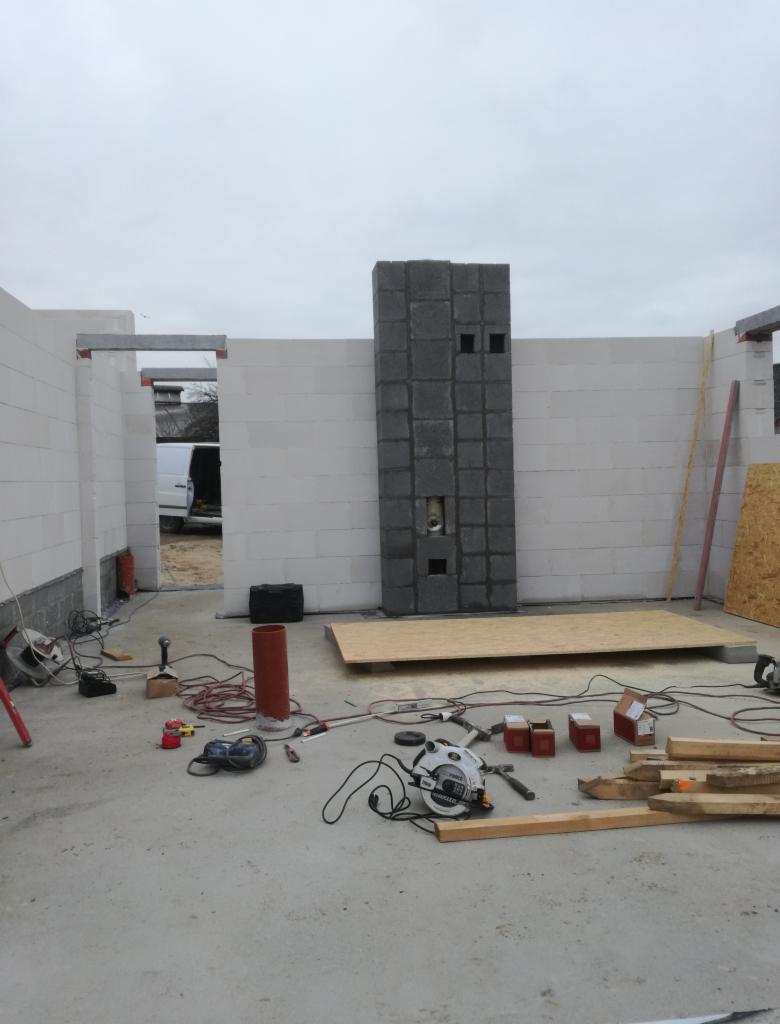 Wnętrze w trakcie budowy: ściany z białych bloczków, komin z szarej kostki, rozłożone narzędzia i materiały budowlane na betonowej podłodze, widoczny samochód dostawczy przez otwór drzwiowy.