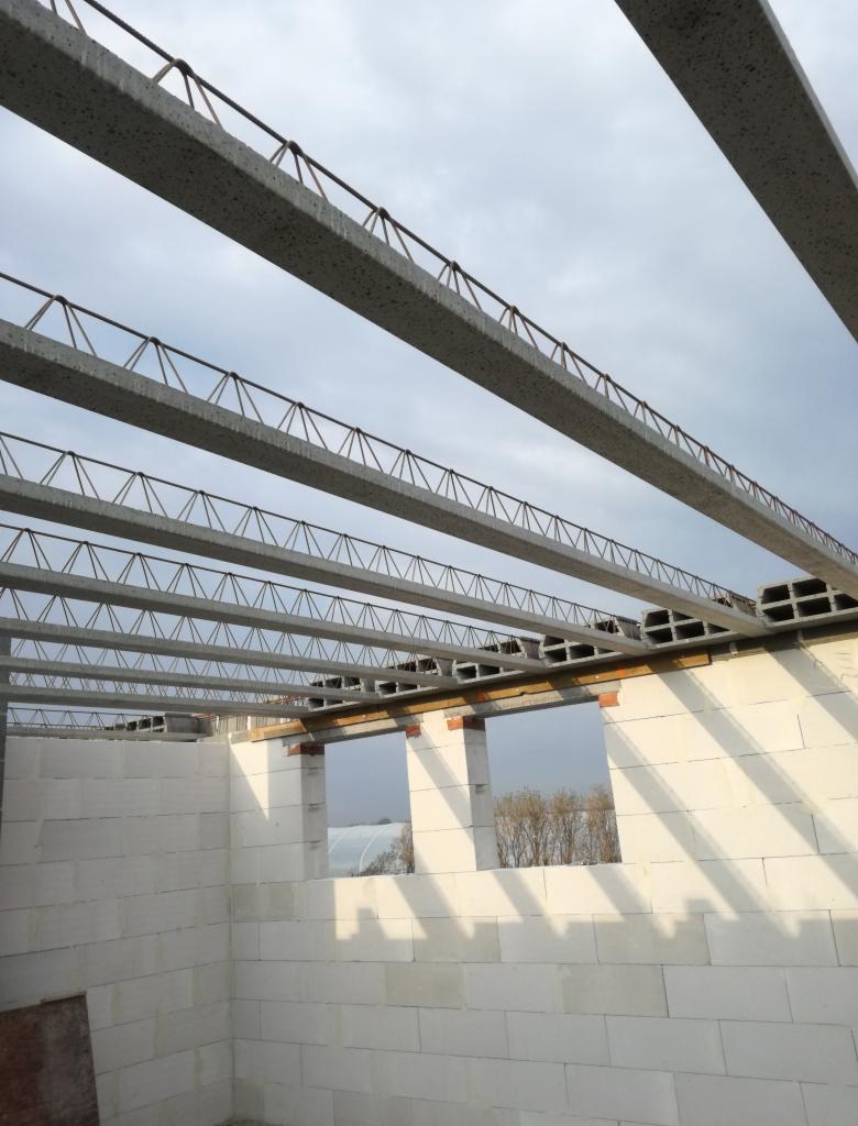 Wznoszenie ścian domu murowanego z betonu komórkowego, widoczne nadproża okienne i prefabrykowane belki stropowe zbrojone stalą, w tle pochmurne niebo.