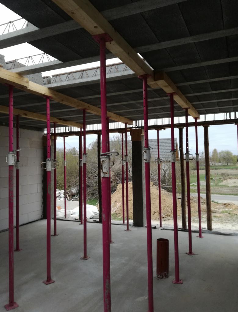 Wnętrze budynku w trakcie budowy z widocznymi podporami stropowymi w kolorze różowym, drewnianymi belkami, pustakami stropowymi oraz fragmentami ścian z białego bloczka.