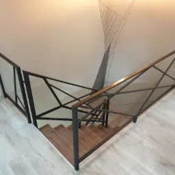 Nowoczesne schody z drewnianymi stopniami i geometryczną, czarną balustradą metalową, widok z góry na tle jasnej ściany z minimalistycznym wzorem.