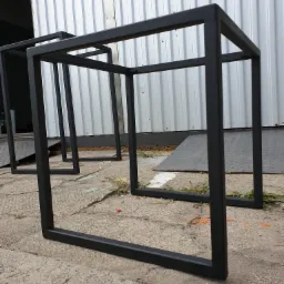 Metalowe ramy w kształcie sześcianu, prawdopodobnie bazy pod kwietniki, pomalowane na czarno, stoją na betonowym podłożu przed blaszakiem.