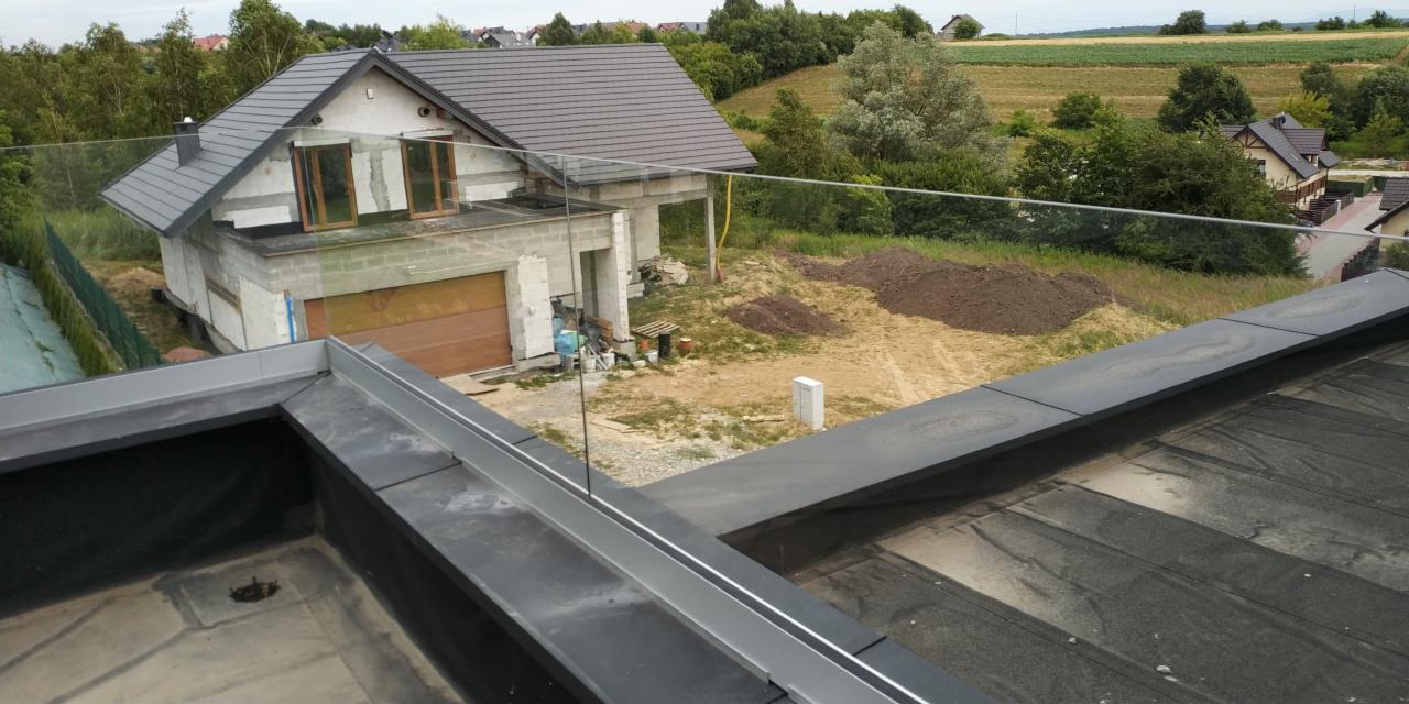 Szklana balustrada na dachu budynku w trakcie budowy, widok z góry na czarne pokrycie dachowe i otoczenie.
