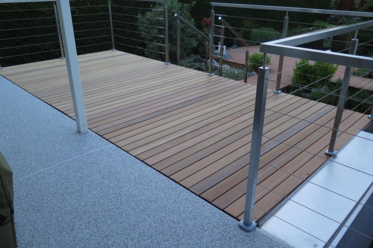 Drewniany taras z balustradą ze stali nierdzewnej, widok z góry, z elementami ogrodu w tle.