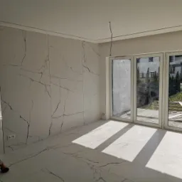 Wnętrze pokoju po remoncie z dużymi, białymi płytami imitującymi marmur na ścianie i podłodze, widoczne przesuwne drzwi balkonowe i drabina oparta o ścianę.