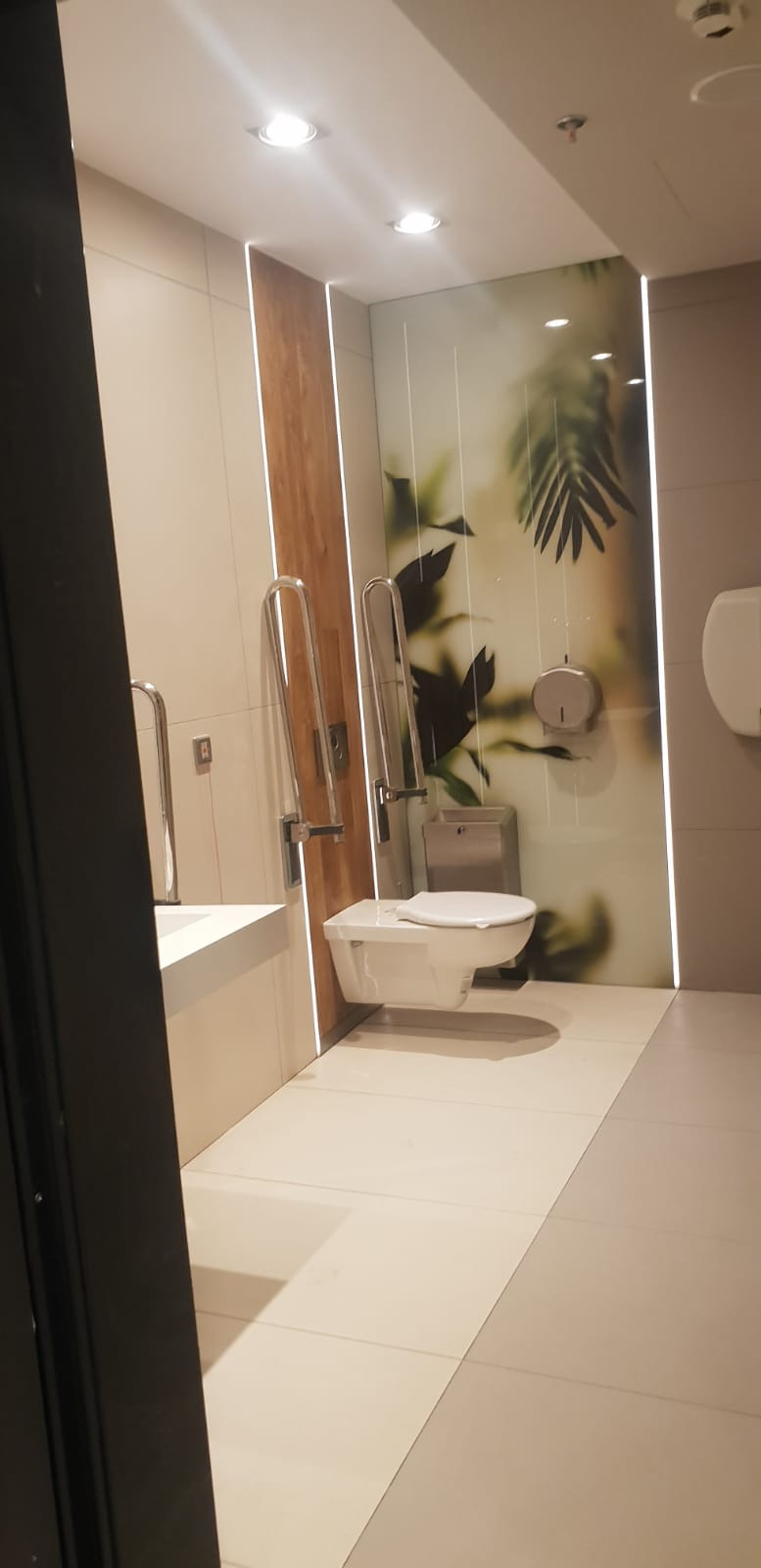 Nowoczesna toaleta w jasnych barwach, wyposażona w uchwyty dla niepełnosprawnych, z dekoracyjną ścianą z motywem roślinnym i podświetleniem LED.