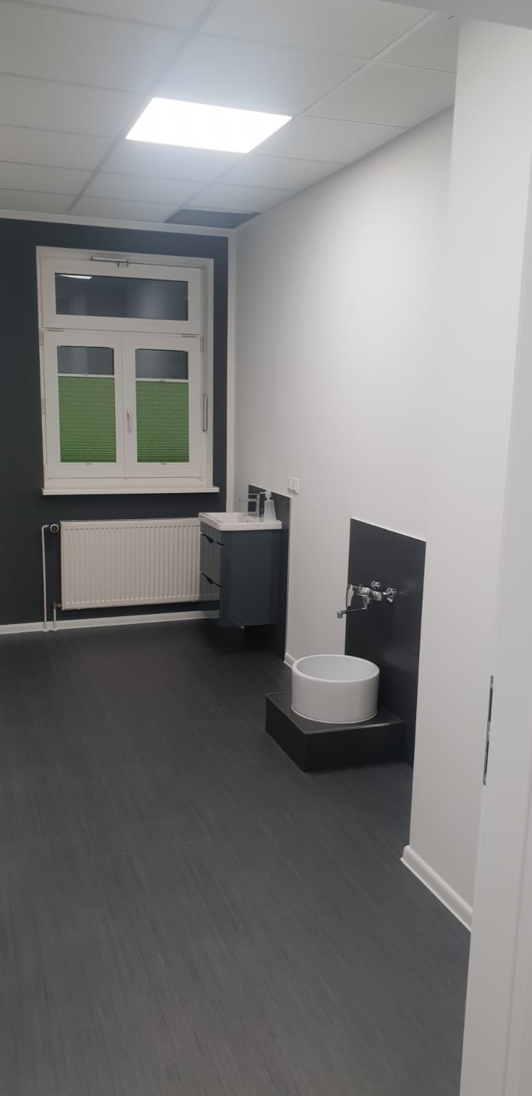 Nowoczesna łazienka z ciemną podłogą, białymi ścianami i oknem z zielonymi roletami, prezentująca umywalkę nablatową na czarnym postumencie oraz szafkę pod umywalkę w podobnej kolorystyce.