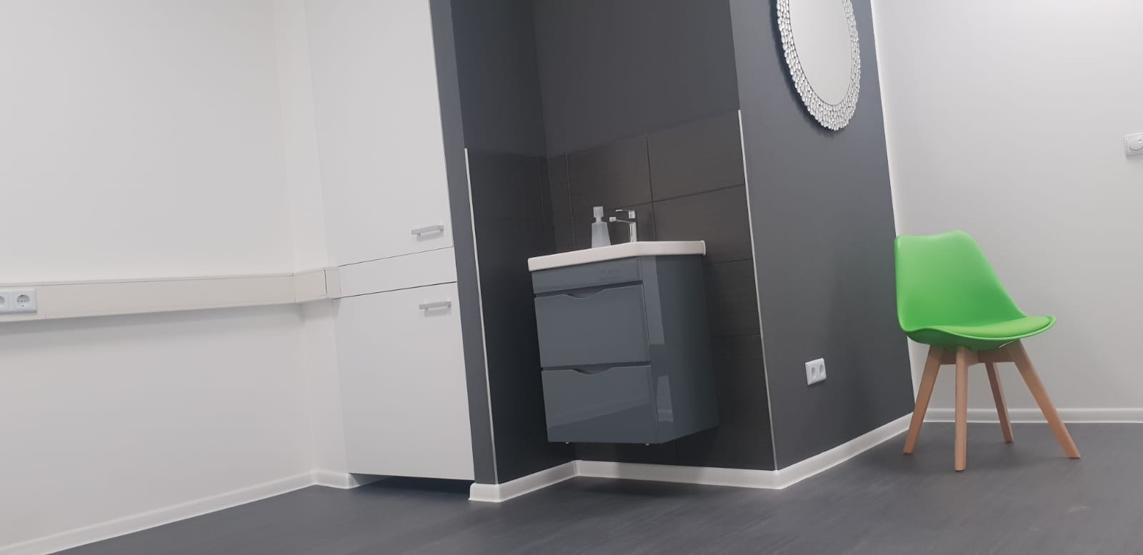 Nowoczesna łazienka z szarą szafką pod umywalką, okrągłym lustrem z dekoracyjną ramą i jasnozielonym krzesłem na drewnianych nogach, na tle białych i szarych ścian.