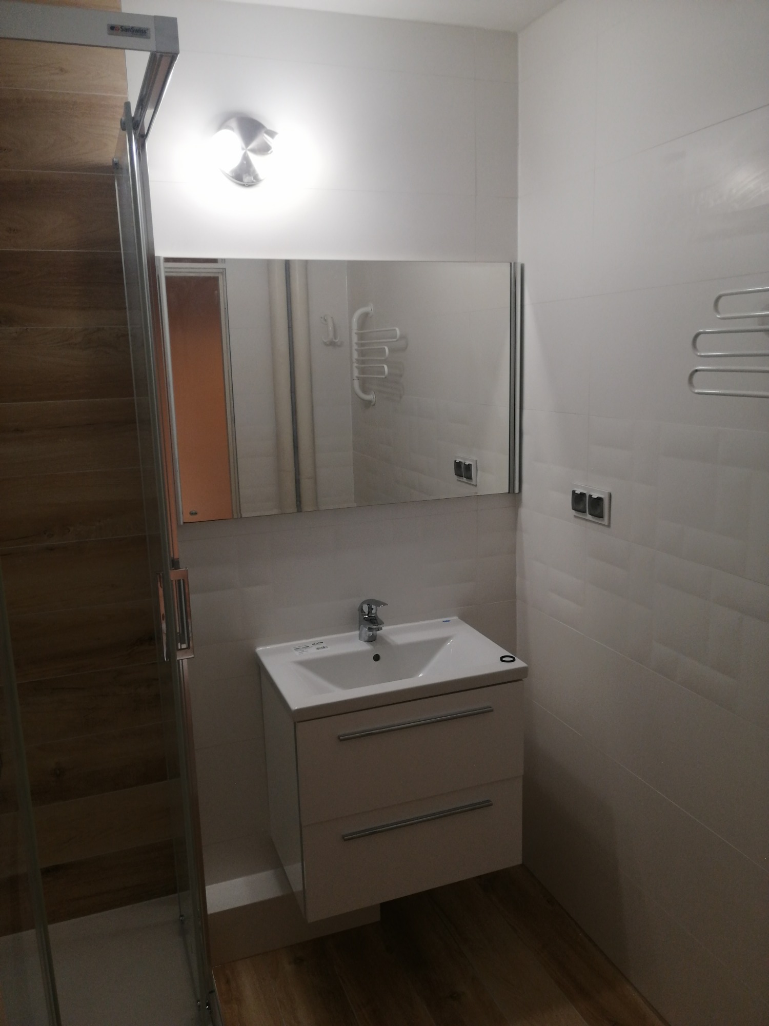 Umywalka z szafką w wyremontowanej łazience, widoczna kabina prysznicowa z drewnopodobnymi płytkami, lustro i grzejnik drabinkowy.