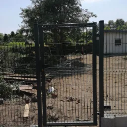 Fragment nowo zamontowanego, czarnego ogrodzenia panelowego z furtką, widoczne betonowe podmurówki i teren po budowie, w tle drzewo iglaste.