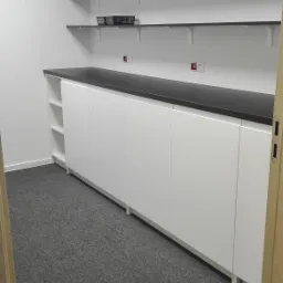 Białe szafki z czarnym blatem w biurze, nad nimi półki z koszykiem, szara wykładzina dywanowa na podłodze.