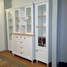 Biały, modułowy mebel z przeszklonymi witrynami i drewnianym blatem, dolne szafki z szufladami i drzwiczkami, wypełniony butelkami, oświetlony lampą ścienną z portretem w tle.