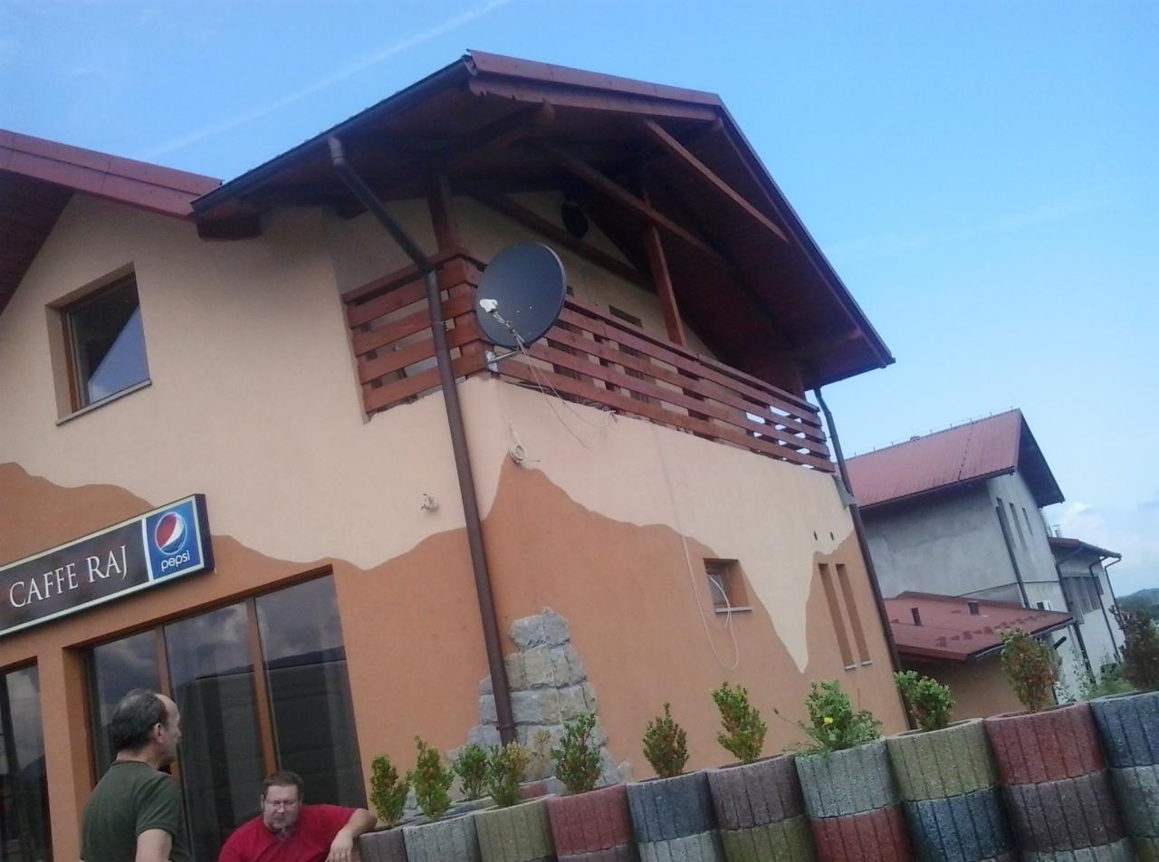 Dwupiętrowy budynek z balkonem i dekoracyjnym malowaniem elewacji w odcieniach beżu i brązu, widoczna antena satelitarna, przed budynkiem donice z roślinami i dwie osoby.