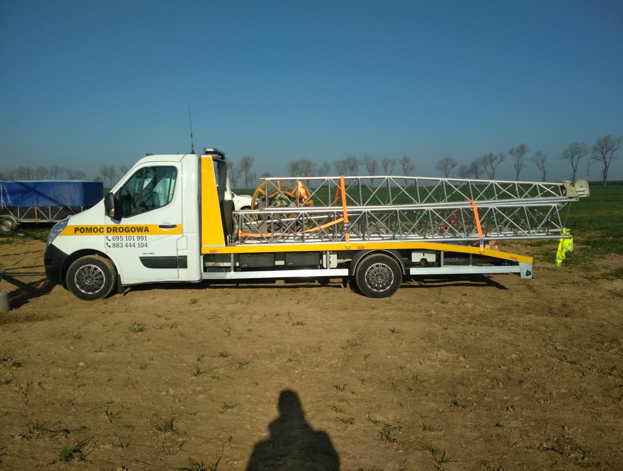 Biała laweta z napisem 'Pomoc Drogowa' przewożąca metalową konstrukcję kratownicową zabezpieczoną pomarańczowymi pasami, na tle zaoranego pola i błękitnego nieba.