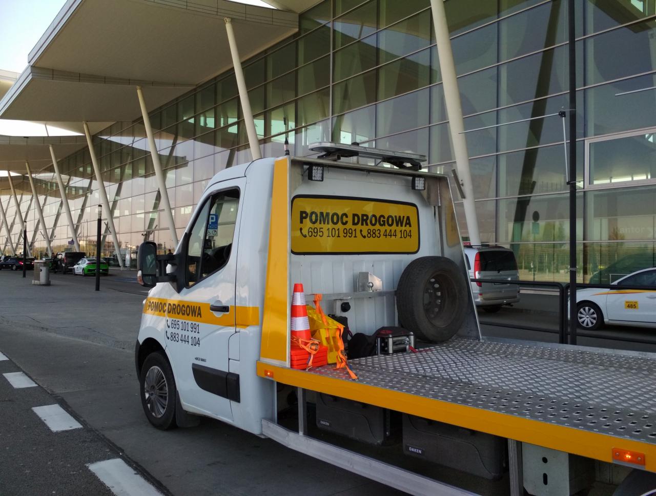 Biała laweta z żółtymi akcentami i napisem 'Pomoc Drogowa' na tle nowoczesnego budynku lotniska, z widocznymi pomarańczowymi pachołkami i kołem zapasowym na platformie.