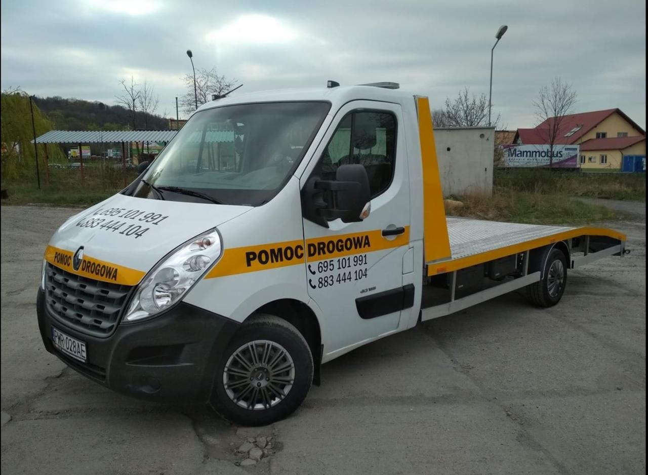 Biała laweta Renault Master z żółtymi akcentami i napisem 'Pomoc Drogowa' oraz numerami telefonów, zaparkowana na szarym asfalcie. W tle widoczny budynek z napisem 'Mammobus' i drzewa bez liści.
