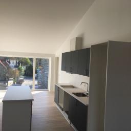 Arton - Jasne, przestronne wnętrze po remoncie z nowymi, szarymi meblami kuchennymi i wyspą na tle dużego okna z widokiem na budynek w rusztowaniach.