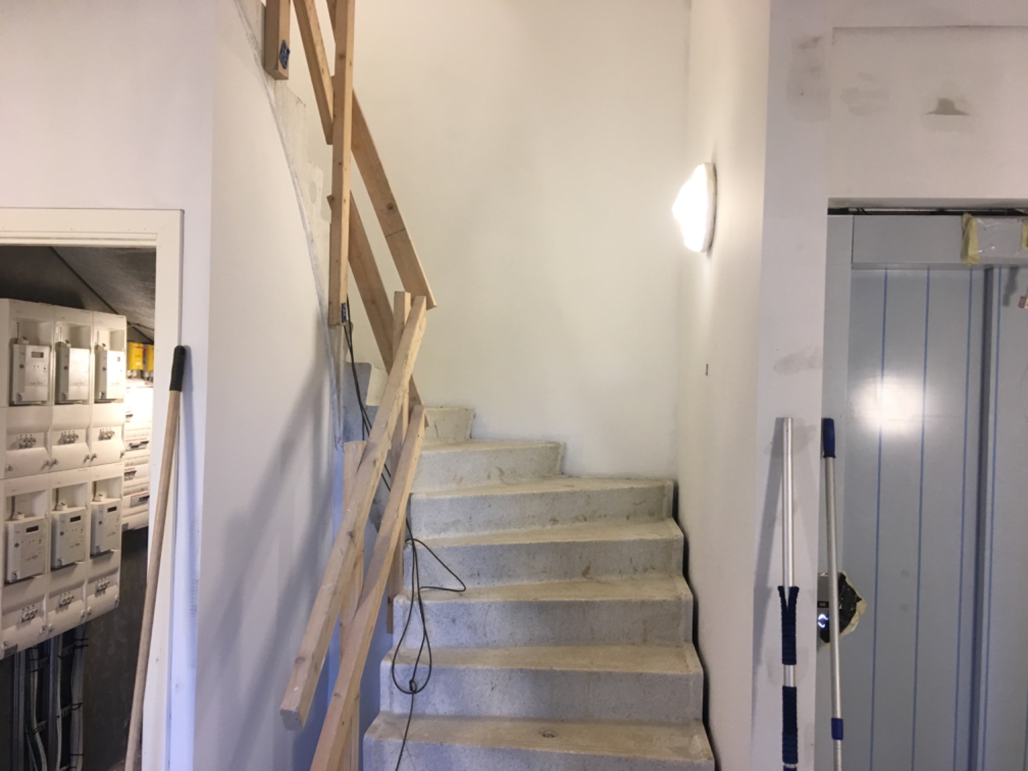 Klatka schodowa w trakcie remontu, widoczne surowe schody, drewniane barierki zabezpieczające, ściany przygotowane do wykończenia, lampa ścienna, mop oparty o ścianę i otwarta szafka z licznikami...