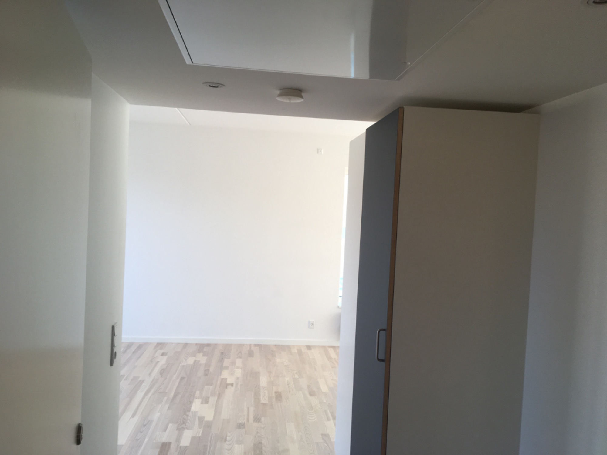 Wnętrze świeżo wykończonego pokoju z jasnymi ścianami, drewnianą podłogą i widocznym włazem na suficie, uchwycone z perspektywy otwartych drzwi.