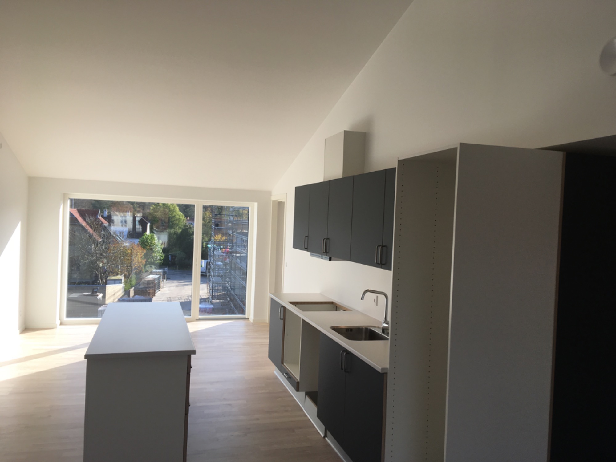 Jasne, przestronne wnętrze po remoncie z nowymi, szarymi meblami kuchennymi i wyspą na tle dużego okna z widokiem na budynek w rusztowaniach.