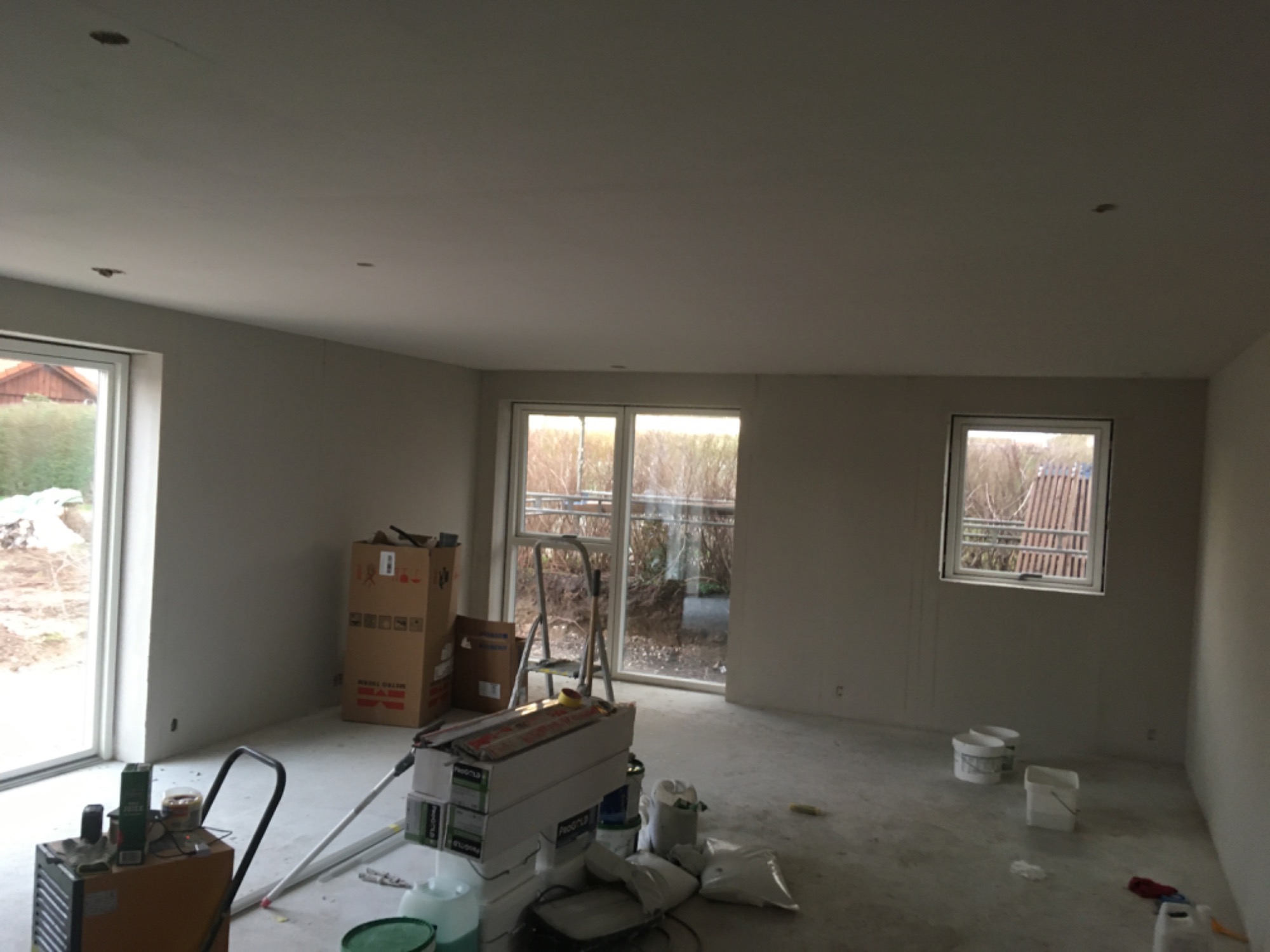 Wnętrze w trakcie remontu: ściany przygotowane pod malowanie, widoczne worki z gładzią, pędzle, drabina, wiaderka i kartony po materiałach budowlanych.