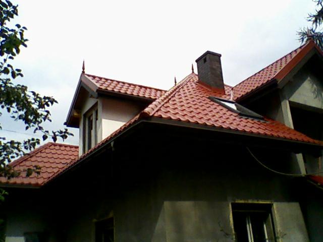 Nowy, czerwony dach z blachodachówki z kominem i oknem dachowym na surowym budynku w trakcie budowy, z widocznymi rynnami i ozdobnymi zakończeniami dachu.