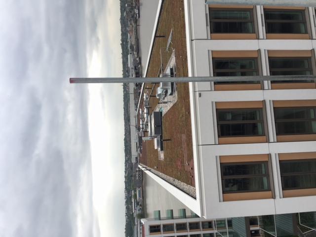 Widok z góry na zielony dach budynku z instalacjami, fragment rusztowania i panoramę miasta w tle, pochmurne niebo.