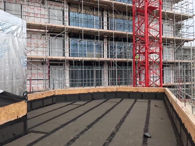 Widok z góry na płaski dach w trakcie hydroizolacji, z widocznymi pasami uszczelniającymi i elementami obróbki blacharskiej przy krawędziach, rusztowania i dźwig w tle.