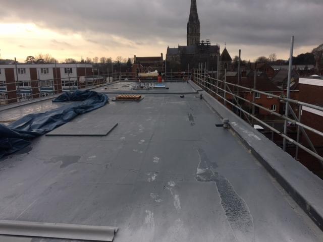 Płaski dach pokryty szarą membraną bitumiczną, widoczne ślady po wodzie, w tle rusztowania, budynki mieszkalne i wieża kościelna pod pochmurnym niebem.